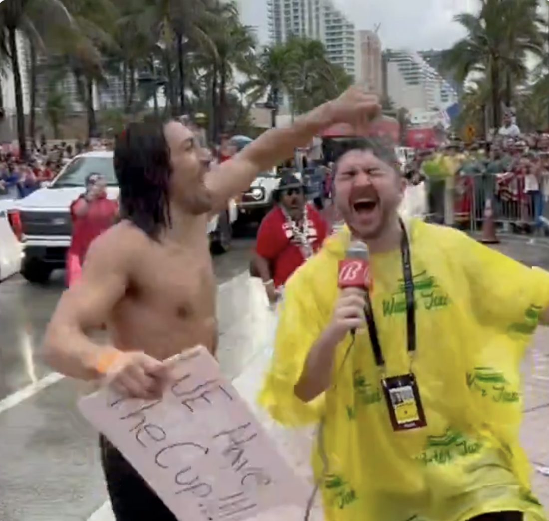 A shirtless fan holding a We Have the Cup sign celebrates with a reporter at the Panthers championship parade